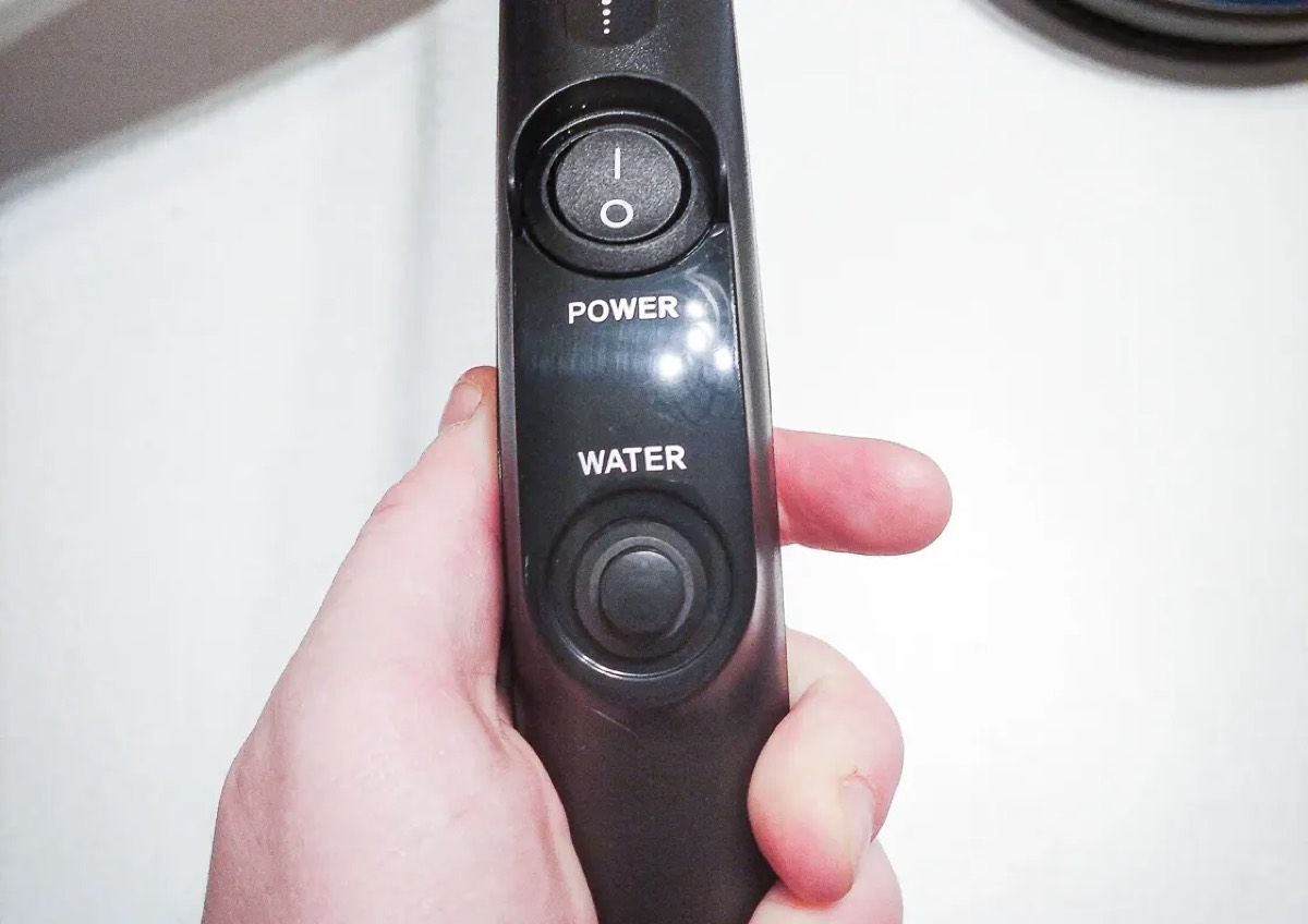 Rączka mopa trzymana w dłoni, widoczny przełącznik POWER i okrągły przycisk WATER